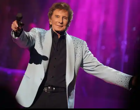 Barry Manilow smiling and pointing toward the audience while holding a microphone