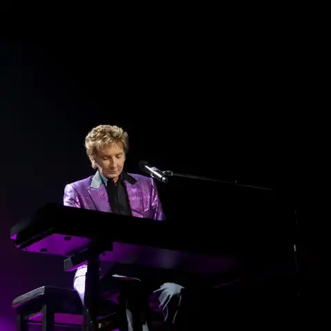 Barry Manilow performing onstage in a light blue rhinestone jacket against purple lighting