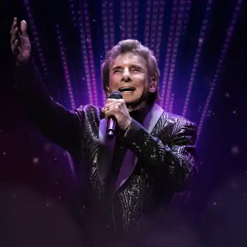 Barry Manilow singing into a handheld microphone on a purple-lit stage