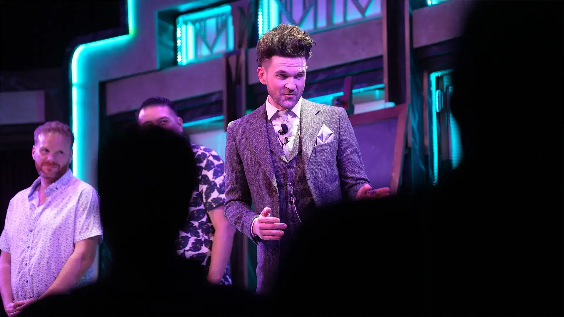 Colin Cloud performing on stage at Harrah's Cabaret in a grey three-piece suit, pointing toward two audience volunteers standing beside him under teal neon Art Deco lighting — Colin Cloud Mastermind Las Vegas