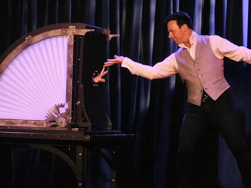 David Goldrake performing a close-up illusion, reaching toward a hand emerging from a steampunk fan-blade box on stage