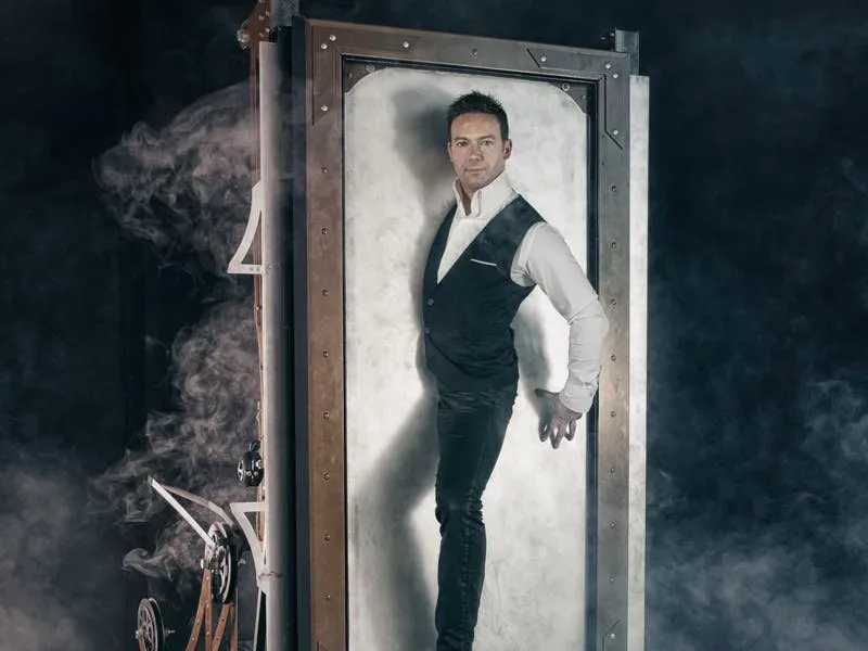 David Goldrake standing inside a riveted metal mirror-box surrounded by smoke at The Magician's Room