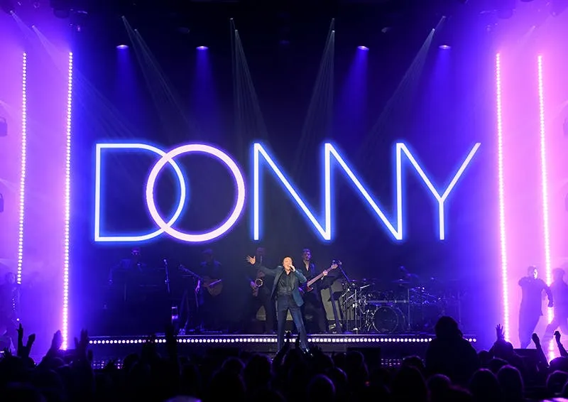 Donny Osmond performing live on stage in front of the glowing DONNY neon sign with his live band