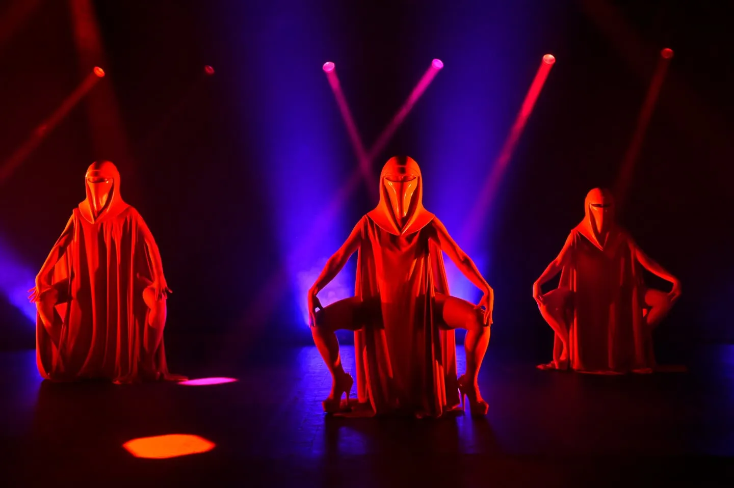 Three red-caped burlesque performers striking a pose on stage under purple and blue lighting
