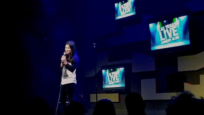 Female comedian performing at Las Vegas LIVE Comedy Club with the show's logo displayed on screens behind her