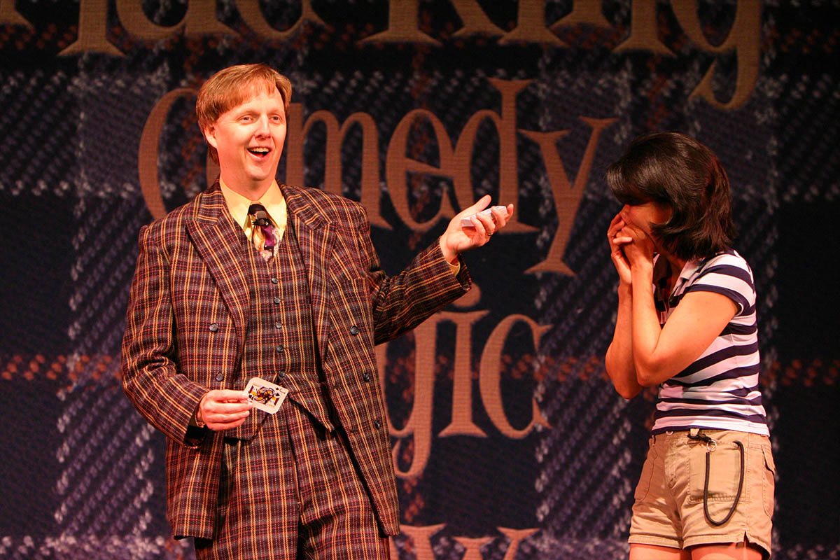Mac King performing a card trick on stage at Thunderland Theater inside Excalibur Hotel Las Vegas as an audience member laughs with delight