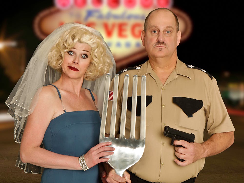 Marriage Can Be Murder cast posing in front of the Welcome to Fabulous Las Vegas sign — bride character in blue dress holding an oversized fork and police officer character, Rio Hotel Las Vegas comedy dinner show