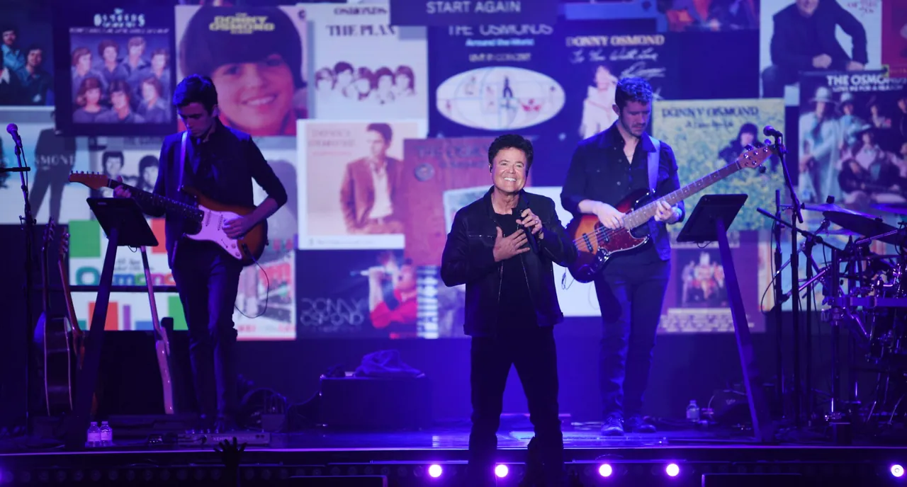 Donny Osmond performing on stage at Harrah's Showroom with dancers and a backdrop of his album covers from across his career