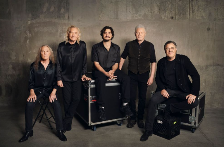 The Eagles — Don Henley, Joe Walsh, Timothy B. Schmit, Vince Gill, and Deacon Frey — posed together in front of a vintage Marshall amplifier stack in a backstage setting