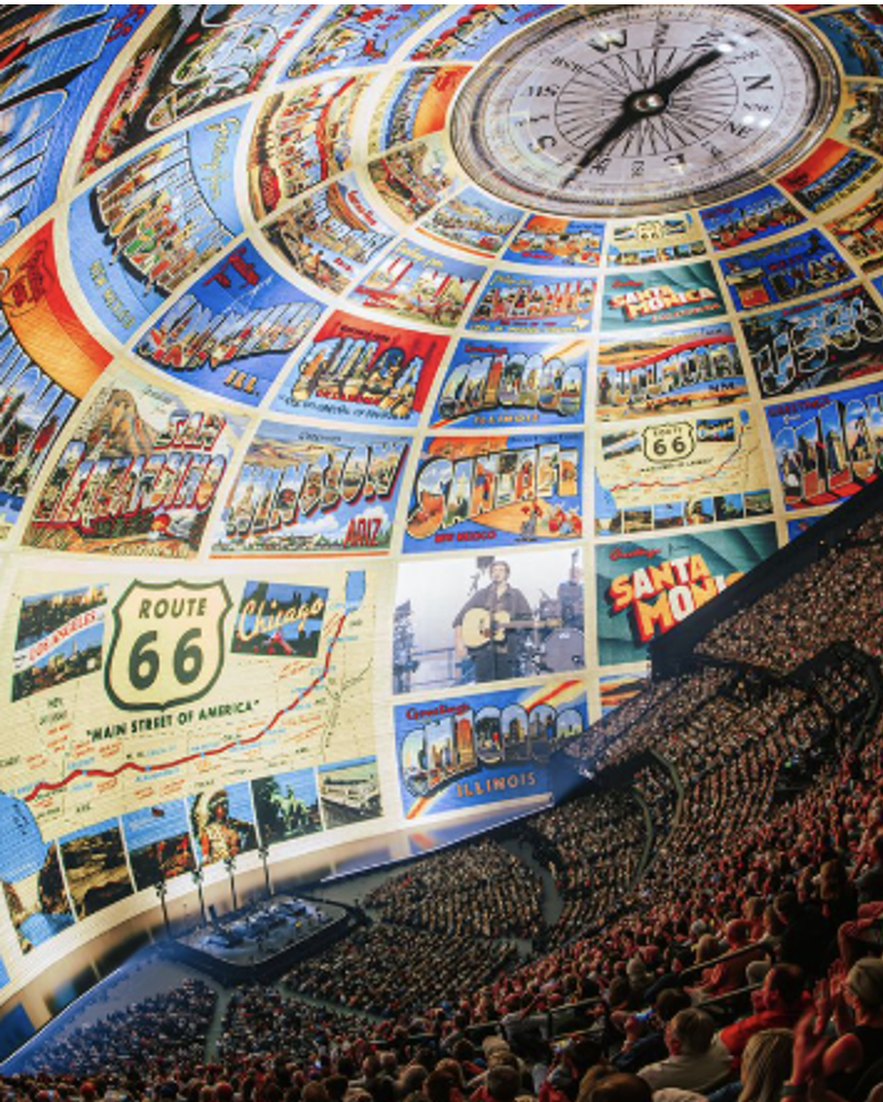 Inside Sphere Las Vegas during an Eagles performance — 18,000 fans look up at the world's largest LED ceiling displaying a massive collage of vintage American Route 66 postcards and Americana imagery, with a lone guitarist performing on stage below