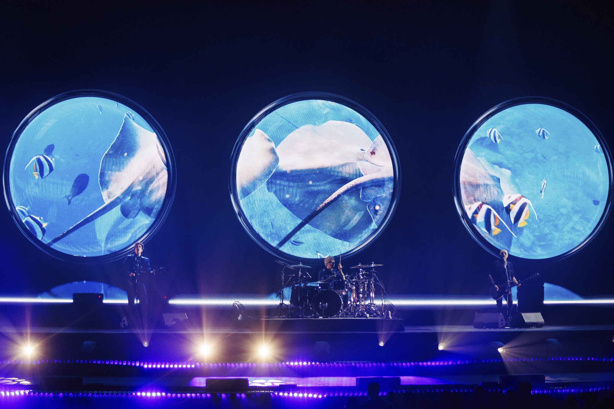 Soda Stereo performing live on the ECOS Tour — three band members on stage beneath three massive circular LED screens displaying underwater ocean imagery, with dramatic blue and purple concert lighting at a packed arena