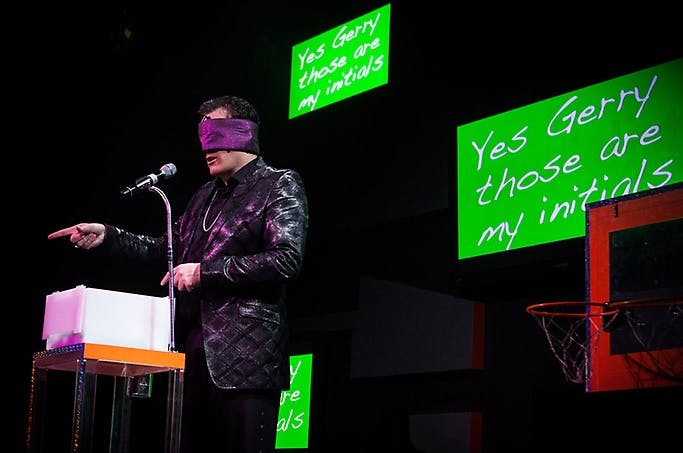 Gerry McCambridge blindfolded on stage pointing toward the audience while large green screens display 'Yes Gerry those are my initials' — The Mentalist mentalism show at Planet Hollywood Las Vegas