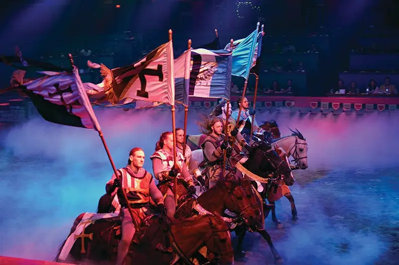 Tournament of Kings — knights on horseback riding through blue smoke carrying flags in the arena