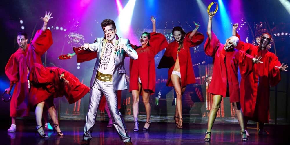 Elvis-inspired zombie performer in a white and gold sequined suit backed by dancers in red robes with green zombie makeup at Zombie Burlesque